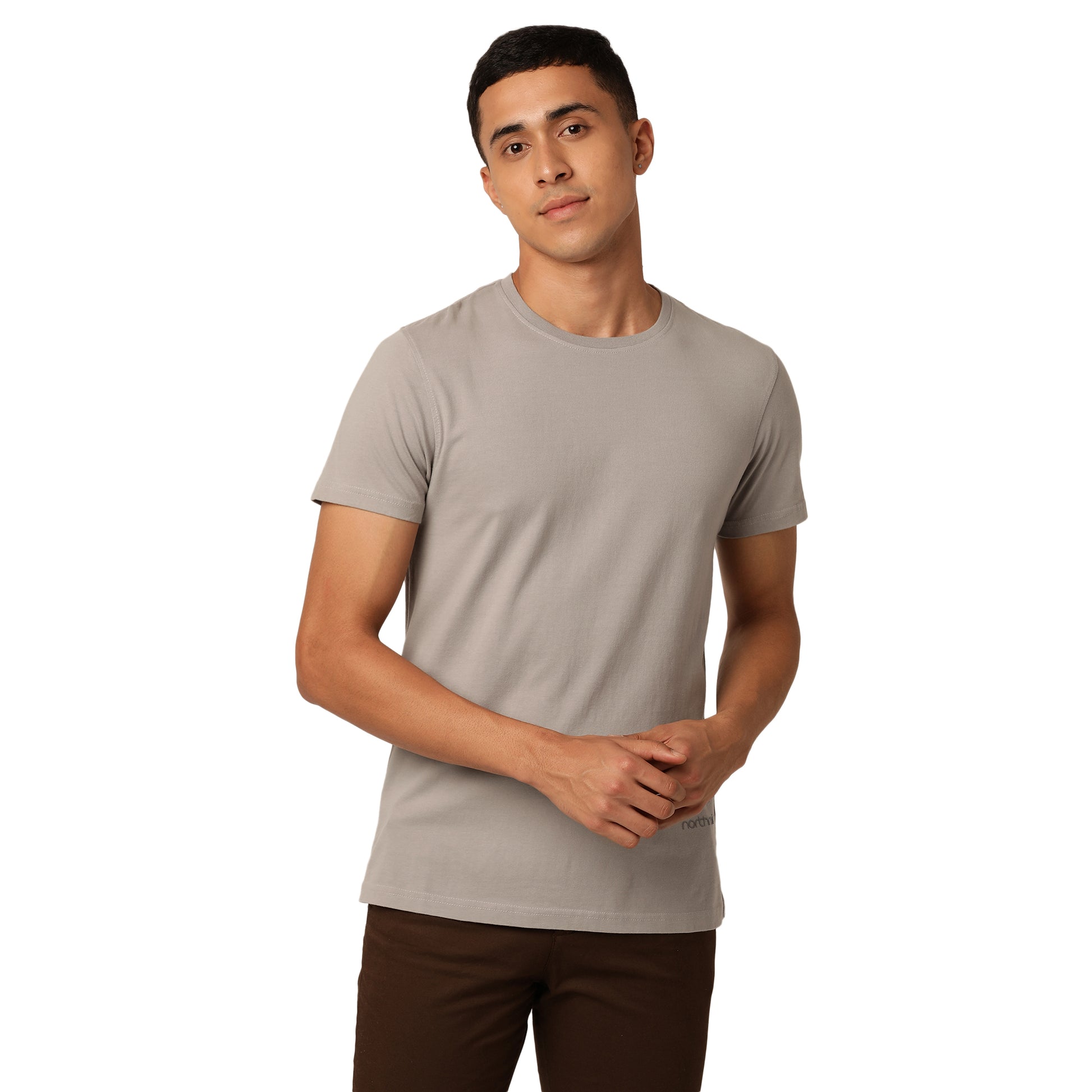 A person with short black hair, wearing a stylish light grey Amity Combo Pack: Organic Crew Neck T-shirt - Grey and Mustard by Northmist and dark brown pants, stands against a plain white background. They have a neutral expression, and their hands are clasped in front of them.