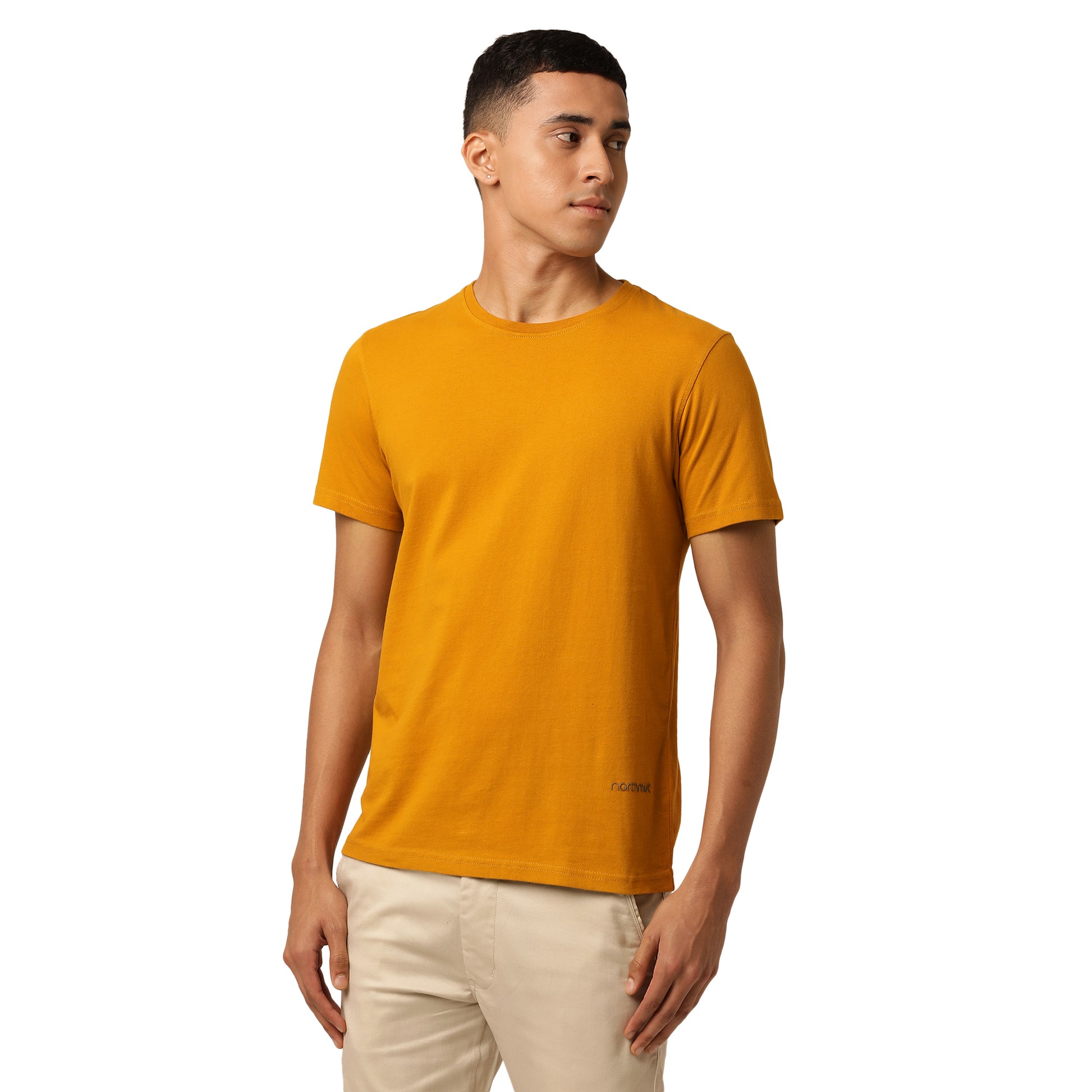 A stylish young man with short dark hair is standing and looking to his right. He is wearing a plain mustard yellow t-shirt made of comfortable organic cotton from the Amity Combo Pack: Organic Crew Neck T-shirts - Grey and Mustard by Northmist, and beige pants. The background is plain white, putting the focus entirely on the man and his outfit.
