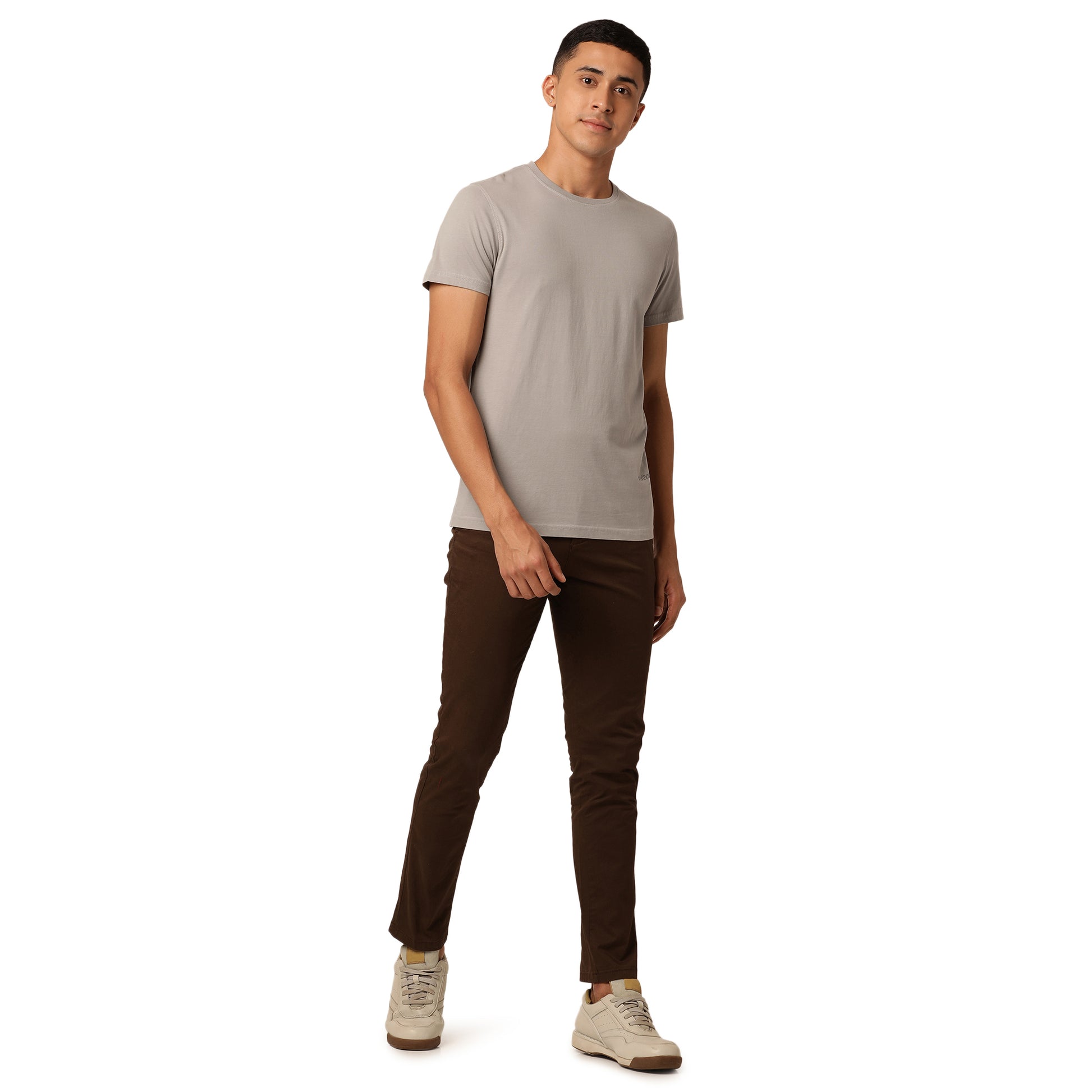 A man stands against a white background, wearing an Amity Combo Pack: Organic Crew Neck T-shirts - Grey and Mustard by Northmist, dark brown pants, and beige sneakers. He is looking slightly to the side with a relaxed expression, with his left hand by his side and his right hand in his pocket.